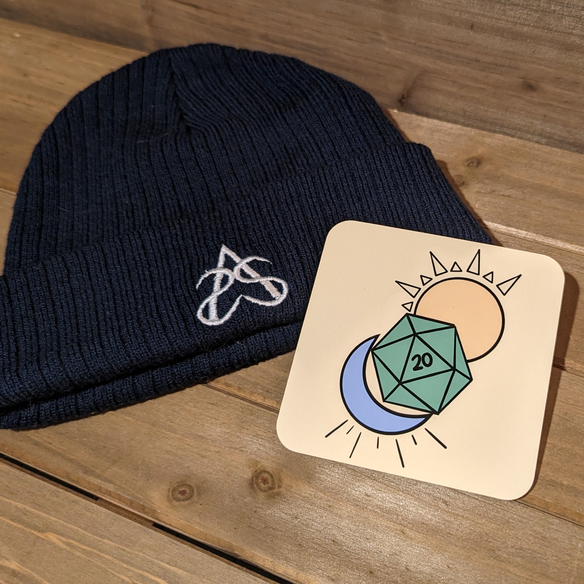 A navy Aras Sivad Studio logo beanie and a coaster with art of a D20, sun, and moon.