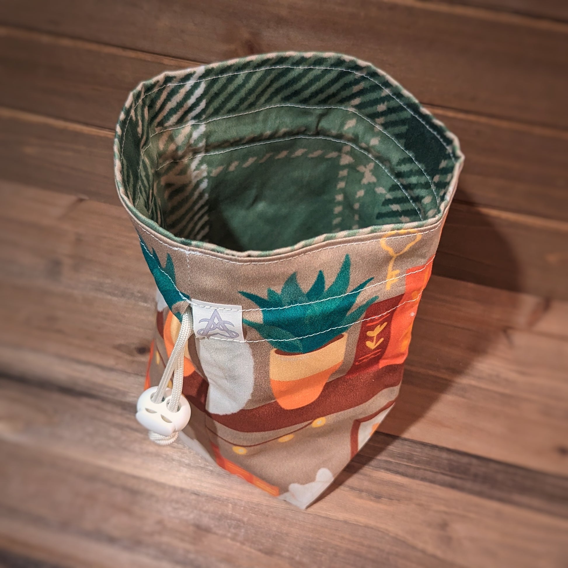 An orange, brown, yellow, and green bookshelf with white cats and potted plants is printed outside a drawstring bag with white stitching and drawstring and a green and cream plaid liner.