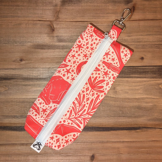 A long, skinny bag has vibrant pink dragon, botanical, and celestial folk art on a soft pink background, a white zipper up the middle, and a keychain clip at the top.