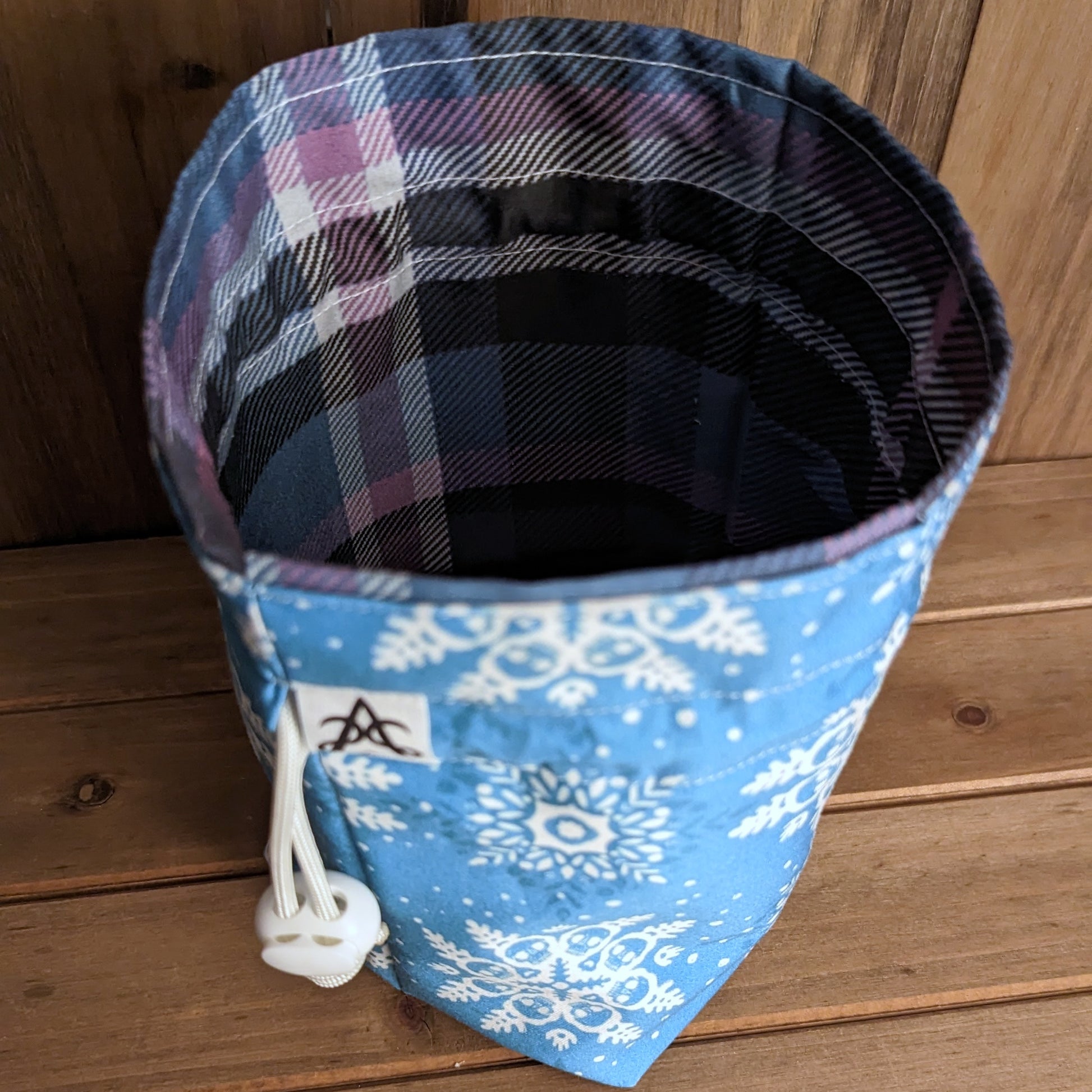 A drawstring bag with a square bottom stands up with a white skull snowflake pattern on light blue outside and a purple and blue plaid liner.
