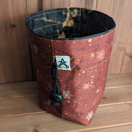 A drawstring bag with a golden snowflake pattern on a red texture print outside, gold stitching, and a liner with gold marbling on black.