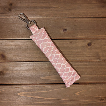 A pen length bag with a keychain clip at the top and a squeeze open on the other side is made with a soft pink doodle style scale pattern.
