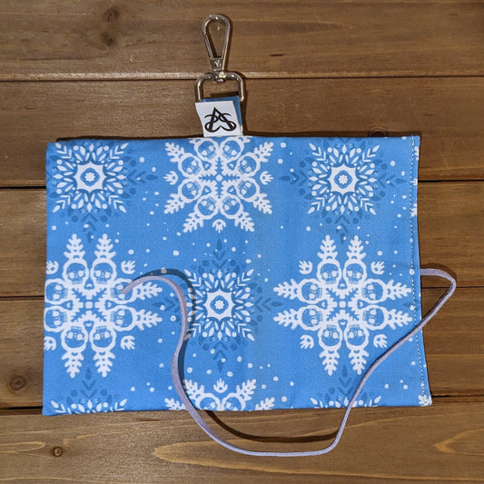 A flat pouch has a white skull filled snowflake pattern on sky blue fabric outside, a fold over flap, and a blue leather cord closure.