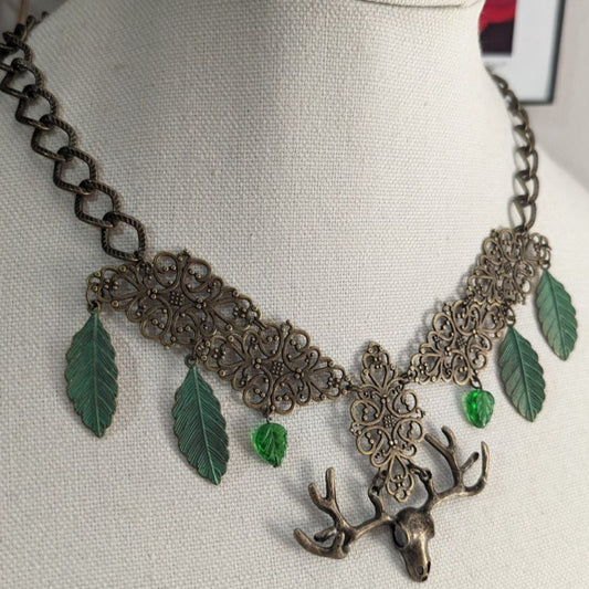 An oxidized brass bib style necklace has four green patina brass leaves, green resin beads, and an antlered skull focal in the middle.
