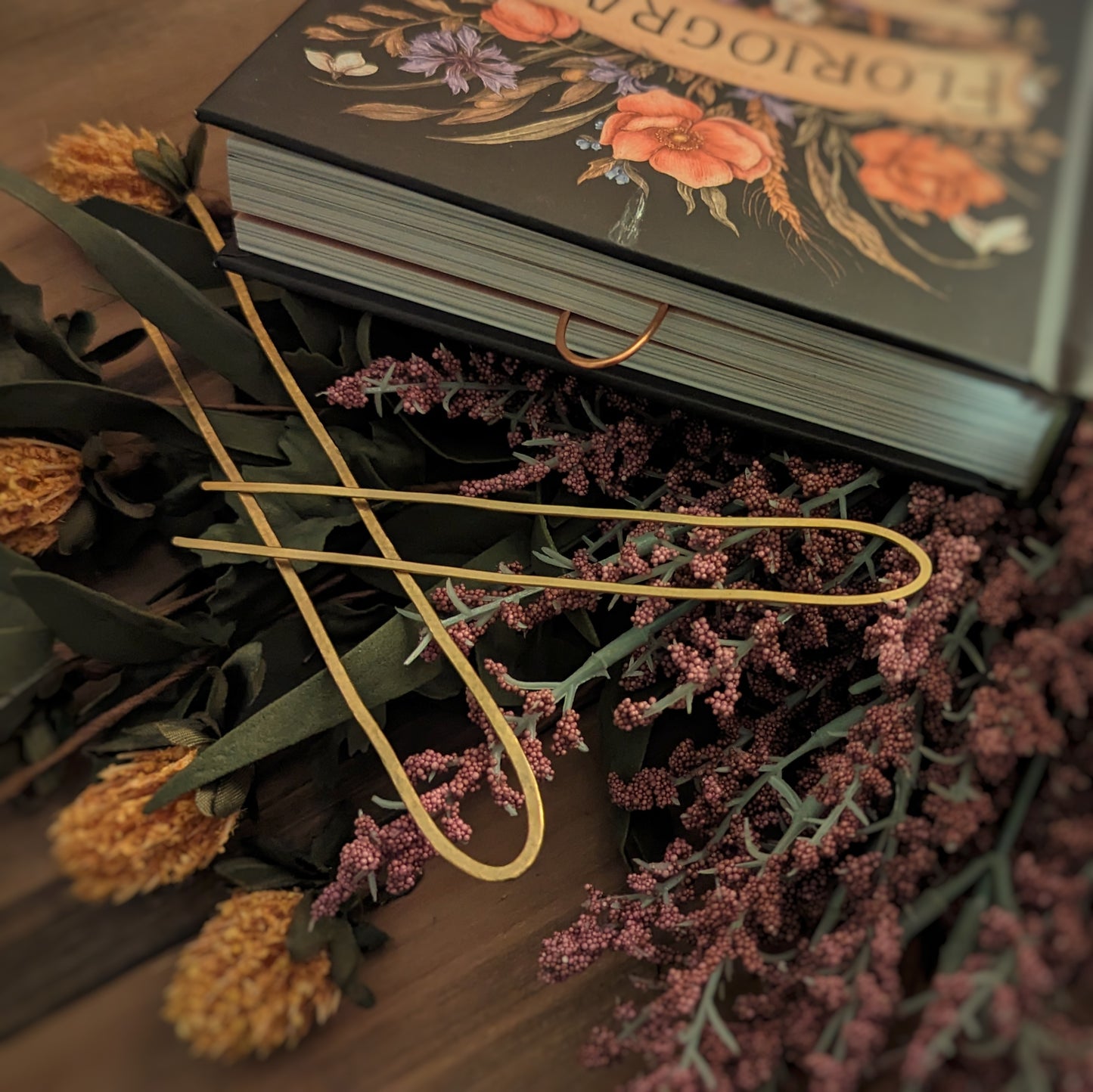 Two hammered brass wire book forks sit on a bed of flowers next to a Floriography book with a pink copper book fork marking two pages.
