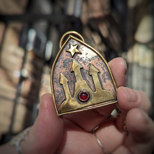 A gold brass castle sits with a red garnet set in the middle of it under a bright gold star, framed by gold wire in front of a patchy pink sky in a pendant held up to the light.