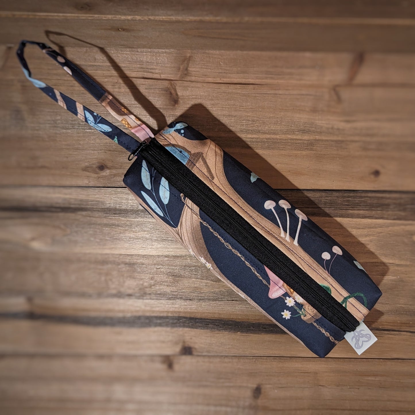 A long skinny pencil case has a fairy village in tree pattern outside, a matching wrist strap, and a black zipper up the middle.