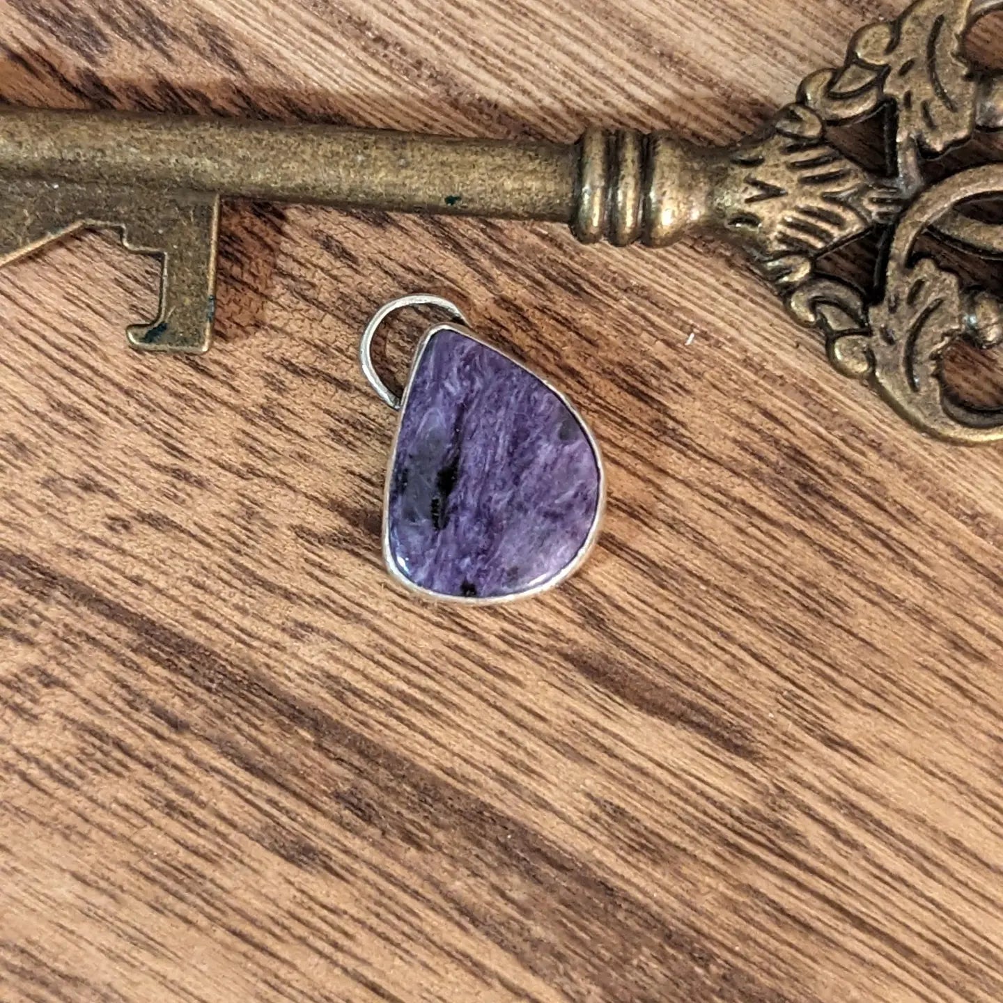 A silver pendant with a solitary purple swirling charoite stone in an almost teardrop shape with one flat side sits with a large brass key.