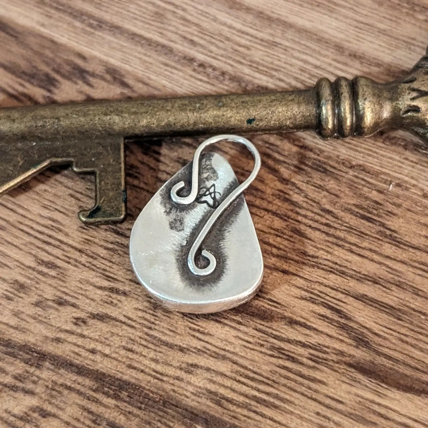 The back of the silver charoite pendant with a slender bail attached to the back in a curling shape and the Aras Sivad logo in a dark patina.