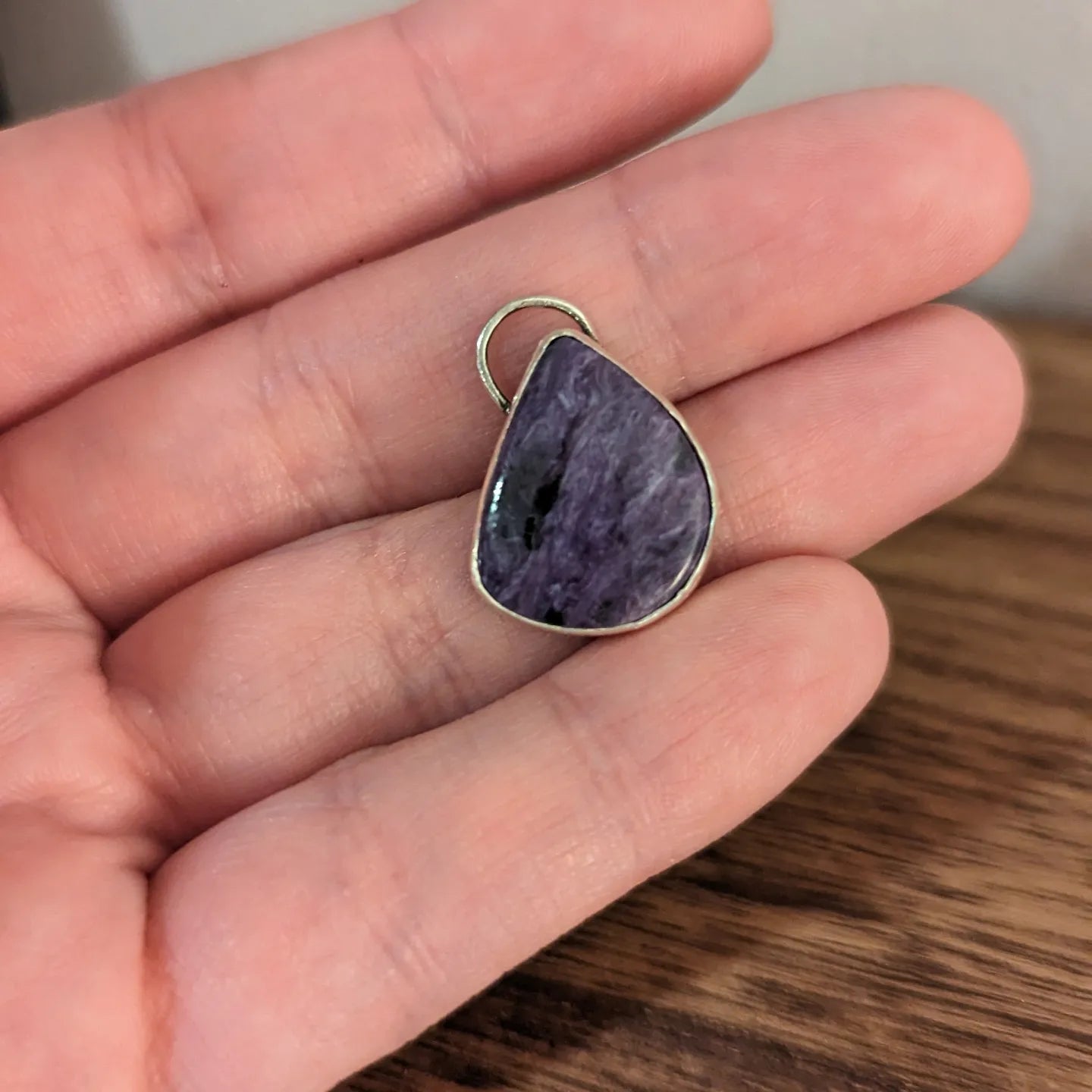 Aras' hand holds a silver pendant with a solitary purple swirling charoite stone in an almost teardrop shape with one flat side.