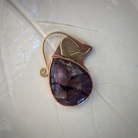 A copper pendant with a teardrop shaped sectioned amethyst cabochon, a gold brass moth resting near the point, and a brass curling bail.