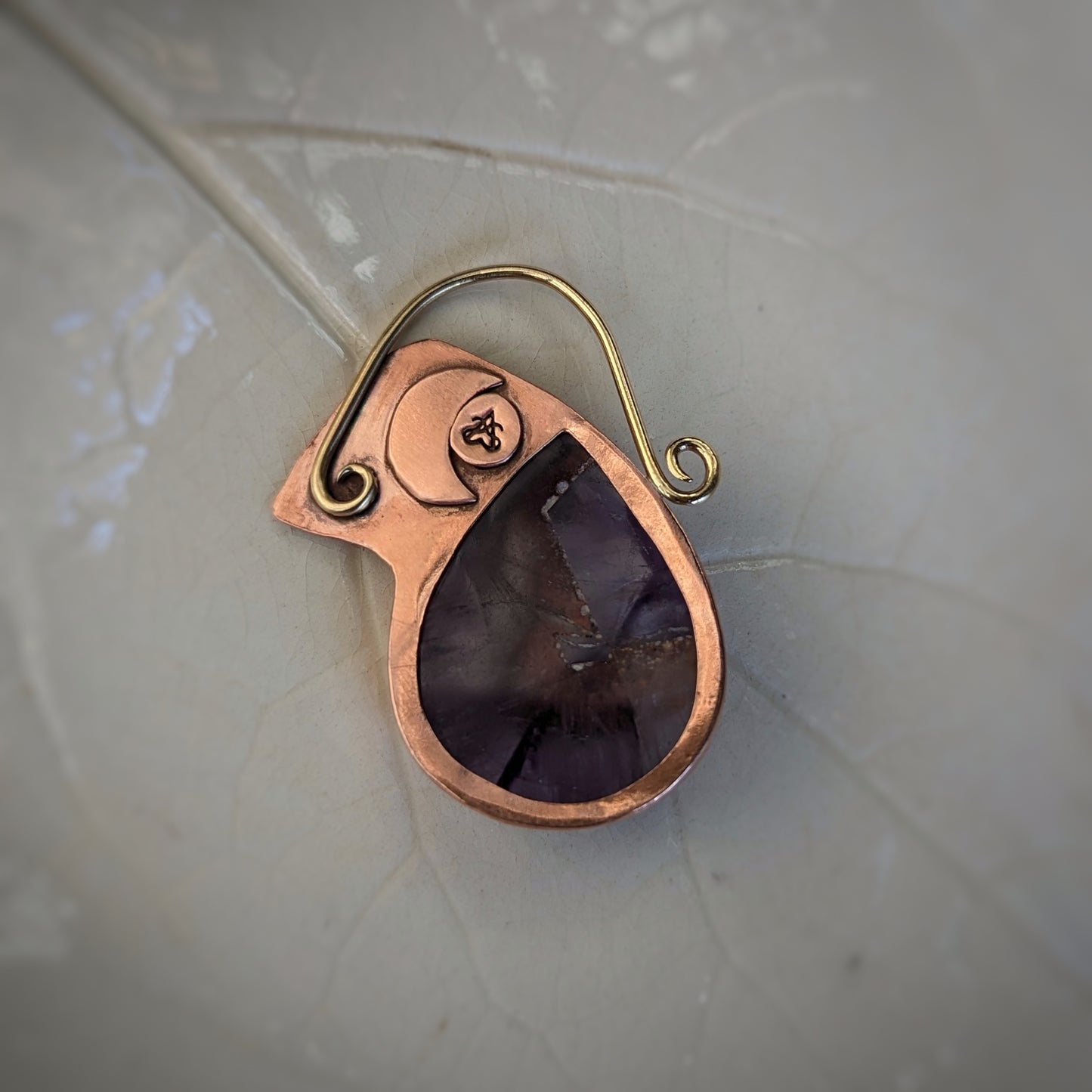 The back of the copper and brass pendant with a teardrop shaped cutout to show off the amethyst with a moon and the Aras Sivad logo on the other side of the moth.