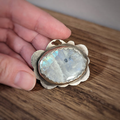 A hand holds a silver cloud pendant with a large oval moonstone in a scalloped edge setting with hammered details on the edges.