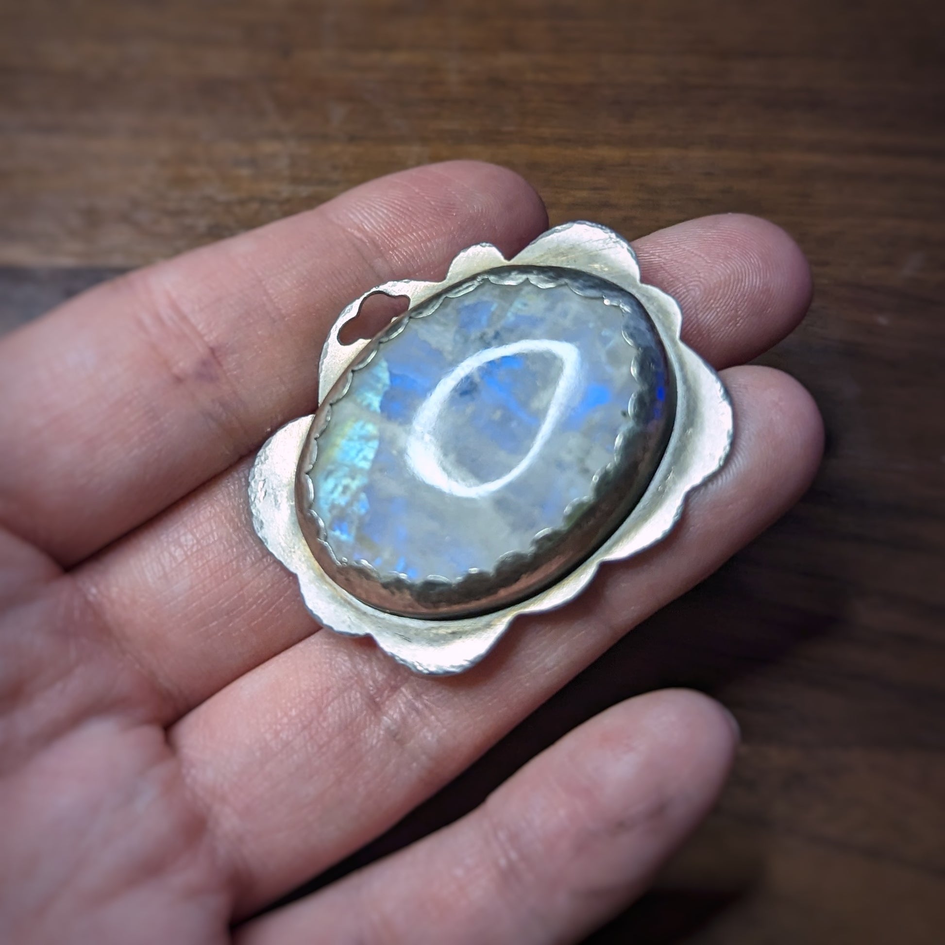 A hand holds a silver cloud pendant with a large oval moonstone in a scalloped edge setting with hammered details on the edges, the moonstone flashing bright blue in the angled light.