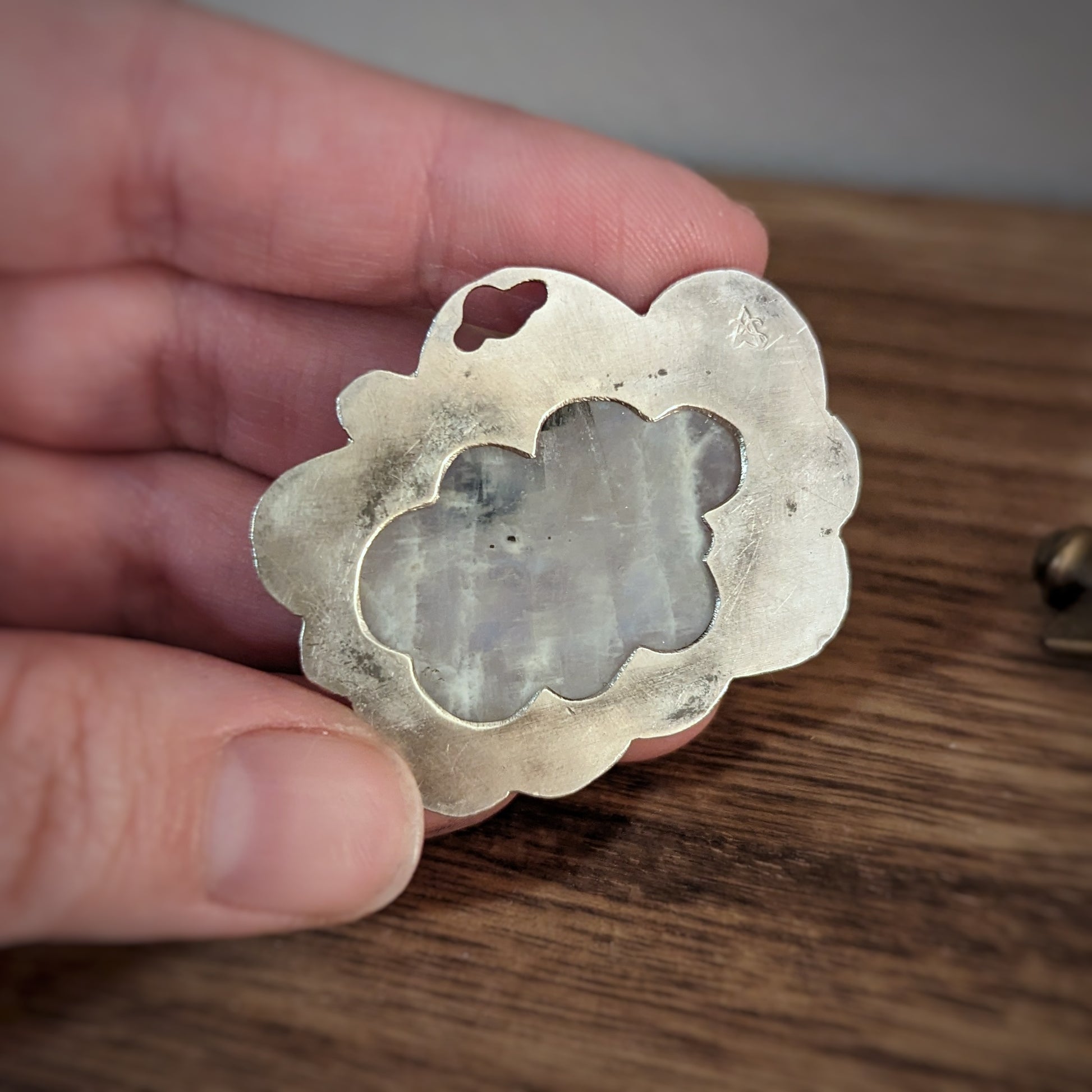 A hand holds a silver cloud pendant with a large oval moonstone in a scalloped edge setting with hammered details on the edges, turned to show a cloud cutout on the back for light to pass through.