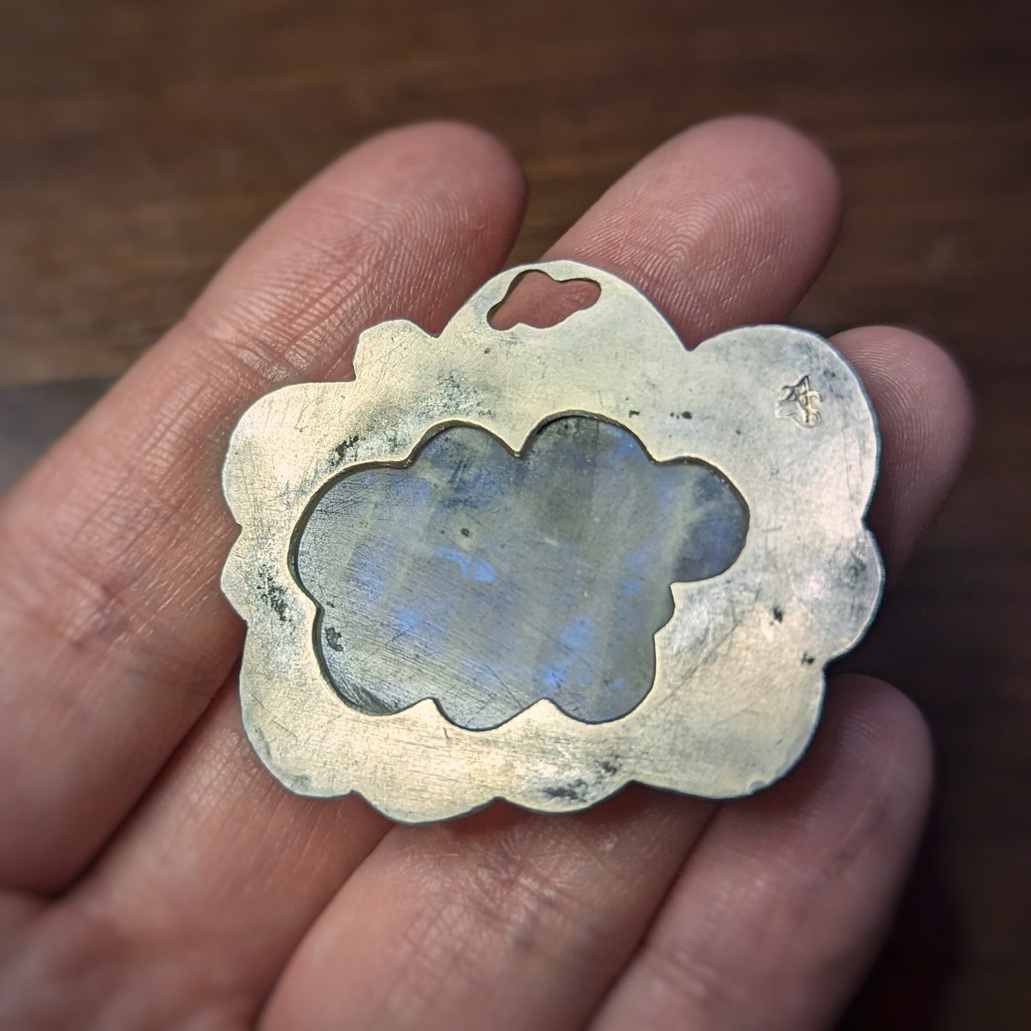 A hand holds a silver cloud pendant with a large oval moonstone in a scalloped edge setting with hammered details on the edges, specks of dark patina in the silver mimicking the tourmaline in the moonstone which flashes blue in the light.