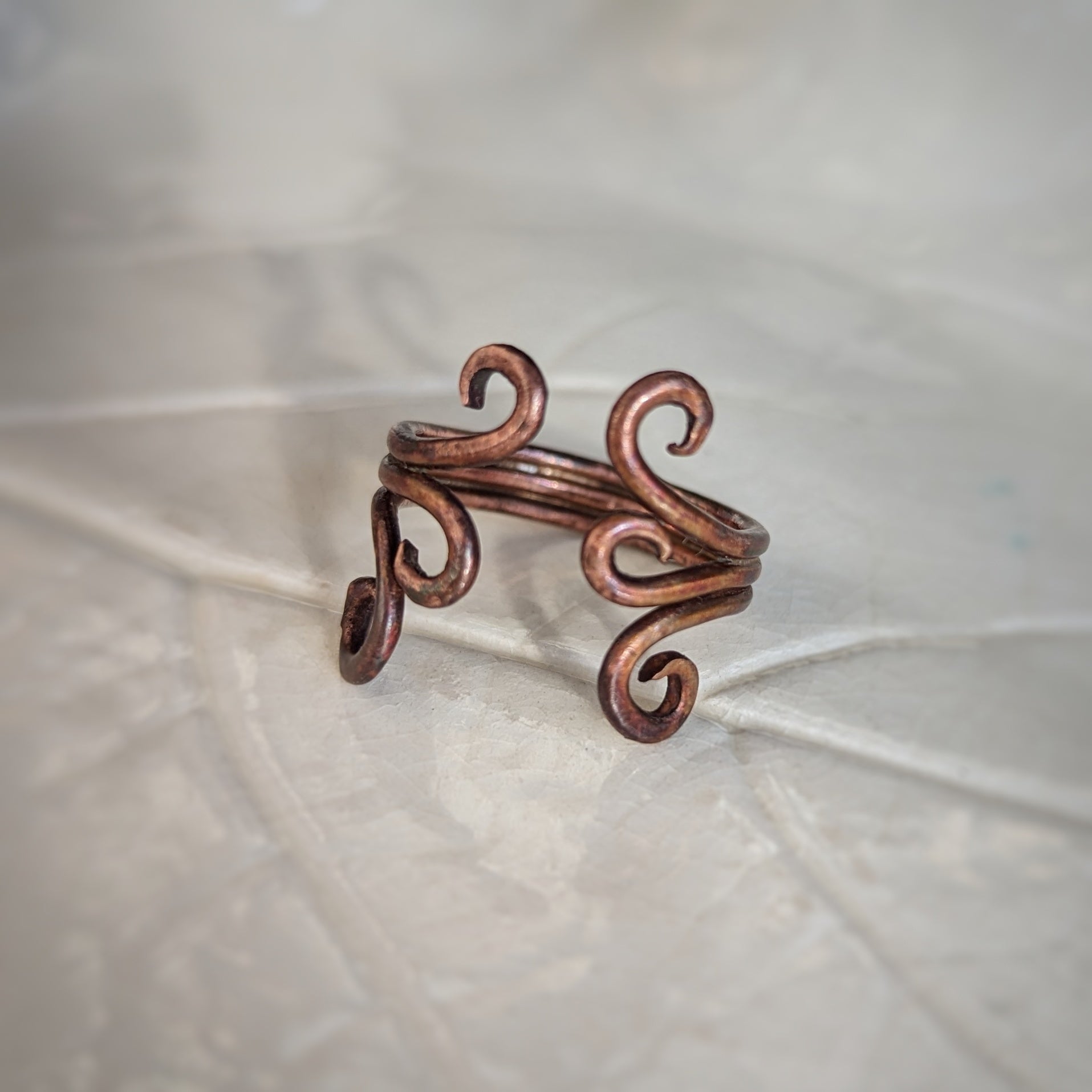 An adjustable copper ring consisting of three wires soldered together, each side ending in graceful swirls has a rainbow appearing patina.