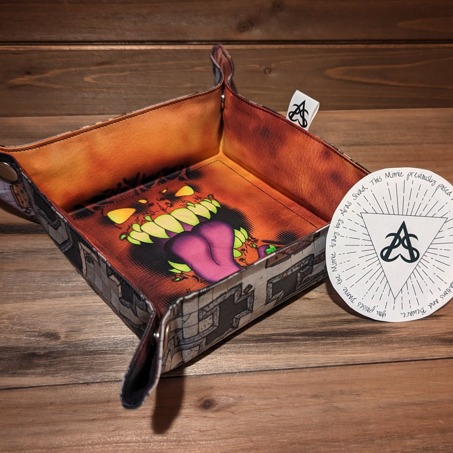A dice tray with a dungeon map exterior and a mimic print liner has a circular certificate of authenticity with the name and a brief story about the mimic.