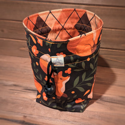 A drawstring bag has curious and sleeping foxes among orange botanicals and vines outside and a peach and brown argyle print liner.