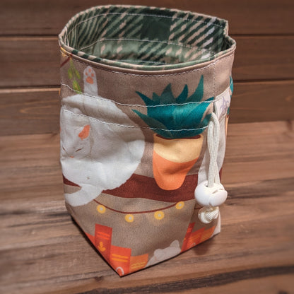 An orange, brown, yellow, and green bookshelf with white cats and potted plants is printed outside a drawstring bag with white stitching and drawstring and a green and cream plaid liner.