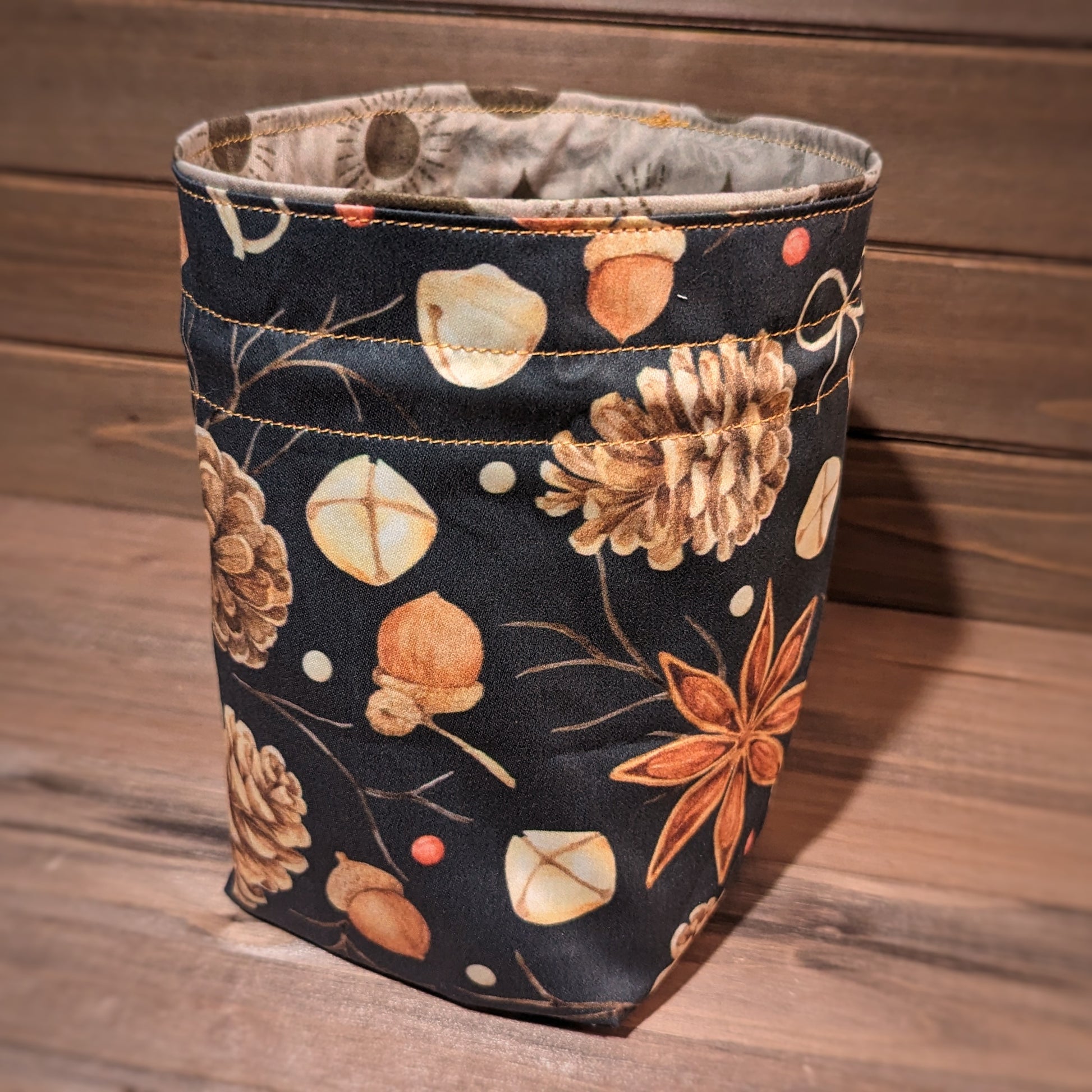 A drawstring bag has pinecone, acorn, star anise, bells, berry, and stick print fabric outside, gold stitching, and sun, moon, and star liner.