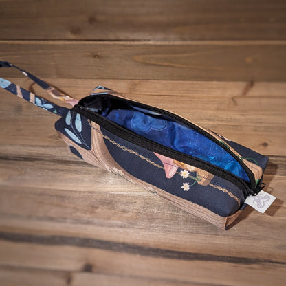 A long skinny pencil case has a fairy village in tree pattern outside, a matching wrist strap, and a black zipper up the middle open to show a blue cloudy design inside.