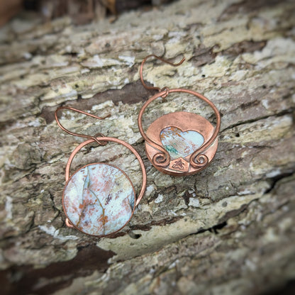 A pair of copper earrings with round petrified wood stones seemingly splattered in fall woodland colors: one earring is turned to show a large heart cut out of the middle, the Aras Sivad logo, and large circular flattened horseshoe shaped bails with swirled ends connected directly to copper hooks.