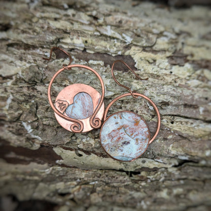 A pair of copper earrings with round petrified wood stones seemingly splattered in fall woodland colors: one earring is turned to show a large heart cut out of the middle, the Aras Sivad logo, and large circular flattened horseshoe shaped bails with swirled ends connected directly to copper hooks.