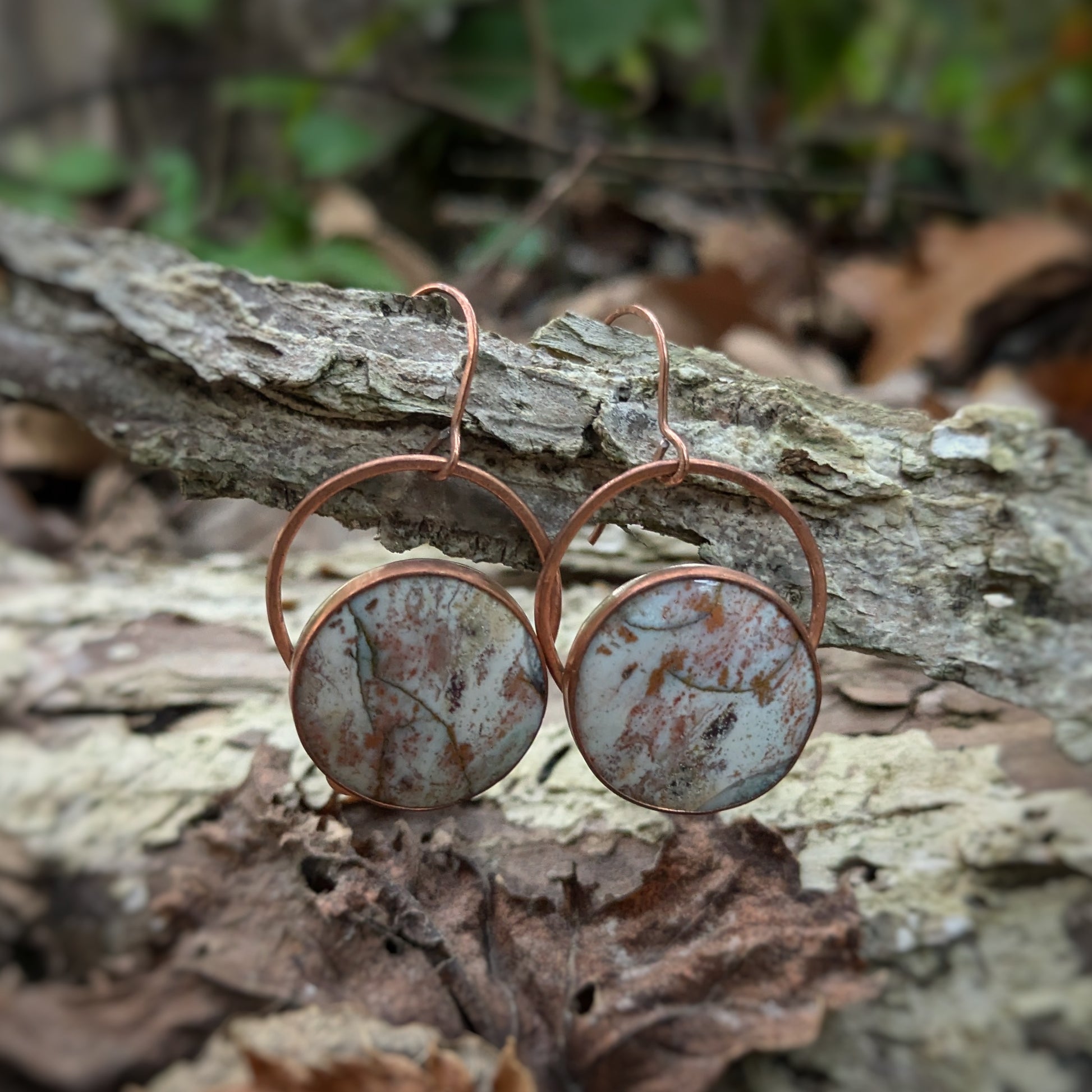 A pair of copper earrings with round petrified wood stones seemingly splattered in fall woodland colors hanging over a silver branch and a bead of leaves in the same color scheme.