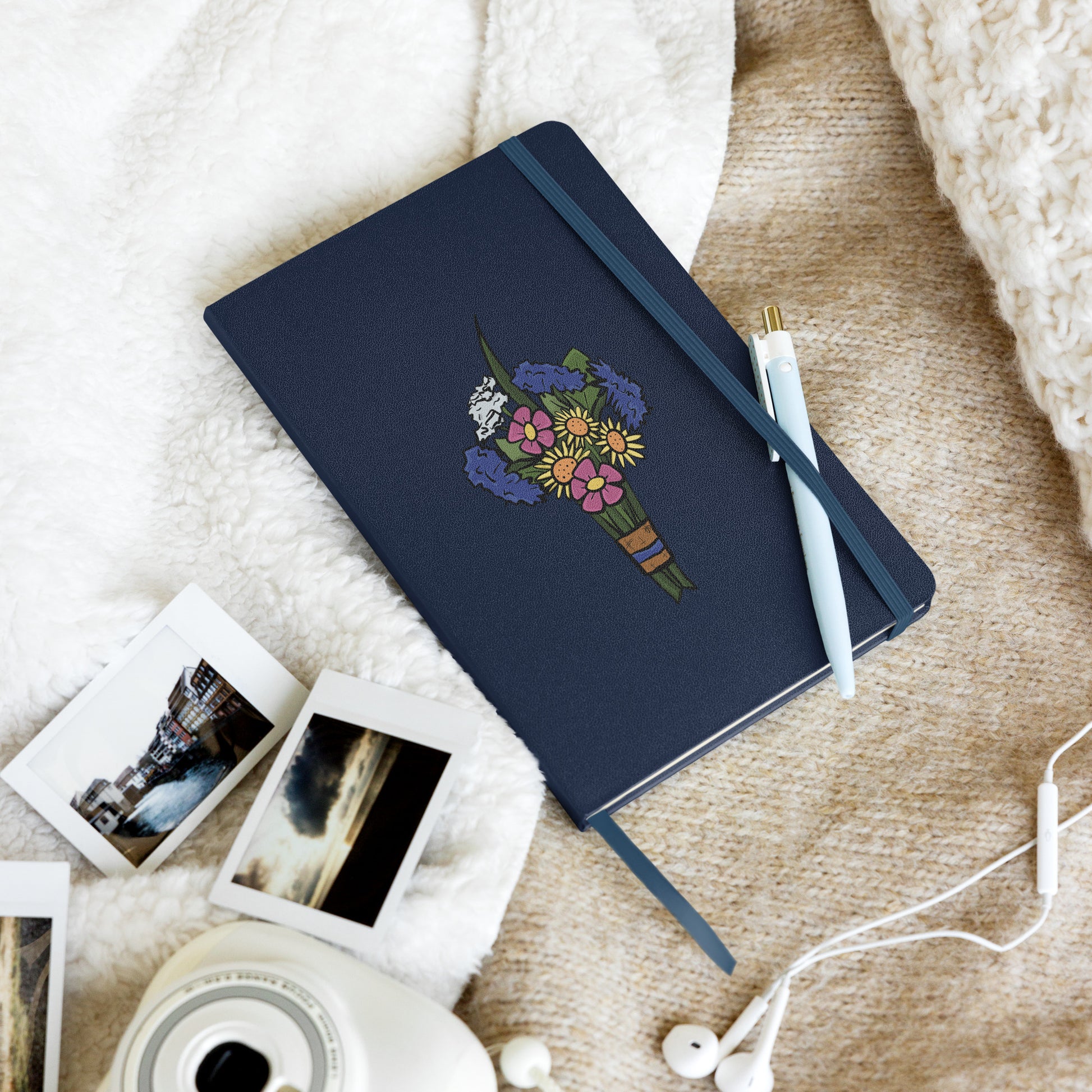 A navy hardcover notebook has a flat colored bouquet of flowers by Aras Sivad on the cover, a ribbon hanging out the bottom, and a pen stuck in its elastic closure.