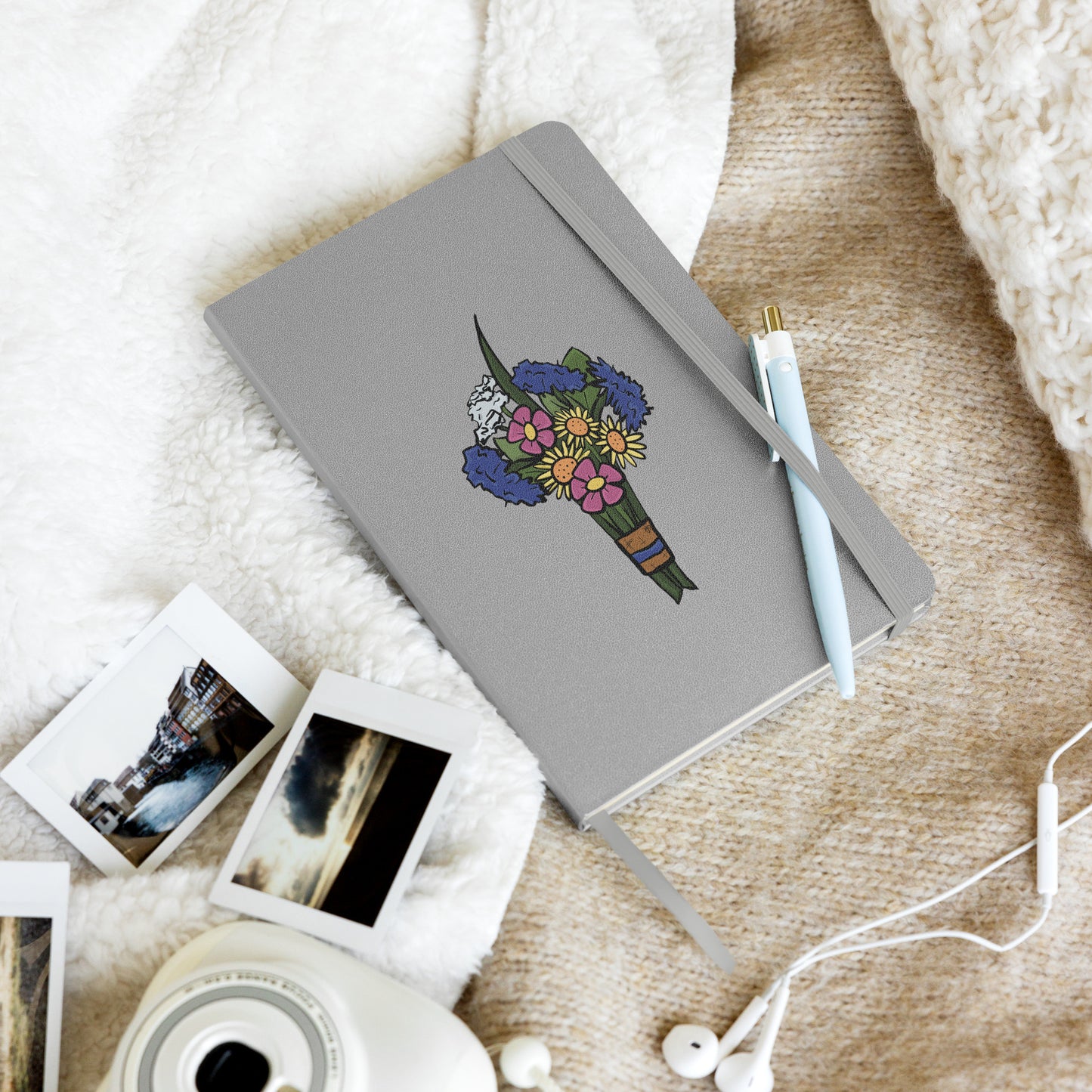 A silver hardcover notebook has a flat colored bouquet of flowers by Aras Sivad on the cover, a ribbon hanging out the bottom, and a pen stuck in its elastic closure.