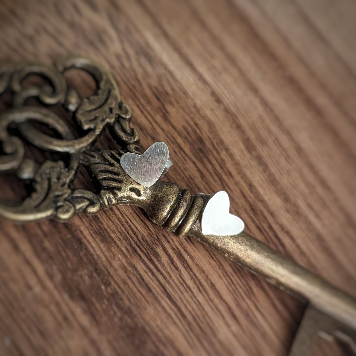 A pair of slightly textured flat silver heart stud earrings sit up against a brass key.