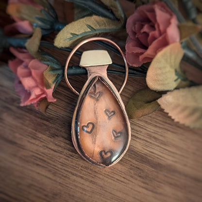 A copper pendant which looks like a potion bottle has stamped hearts under an almost clear rose quartz and a brass cap.