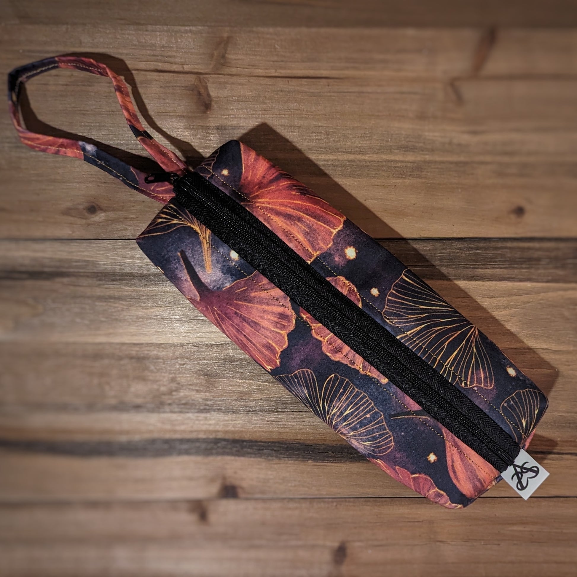 A long skinny pencil case has a gold and pink gingko print outside with matching wrist strap and black zipper down the middle lengthwise.