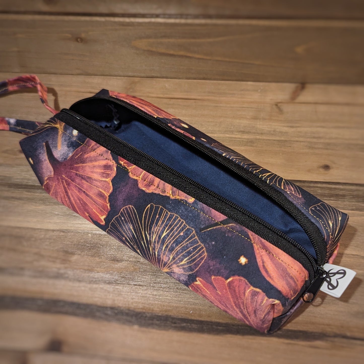 A long skinny pencil case has a gold and pink gingko print outside with matching wrist strap and black zipper down the middle lengthwise is open to show a navy blue tonal liner.