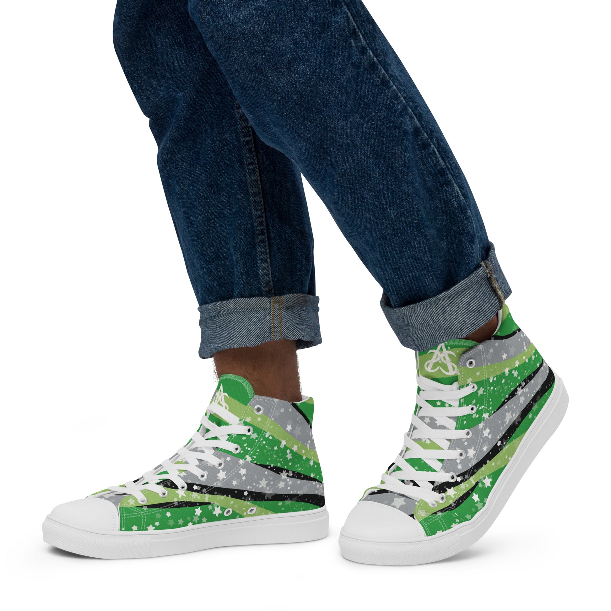 A model wears rolled up jeans and a pair of high-top shoes with ribbons of the greens, grey, and black of the aromantic pride flag coming from the heel and expanding towards the laces with an explosion of stars over it, white accents, and the Aras Sivad Studio logo in white on the tongue.