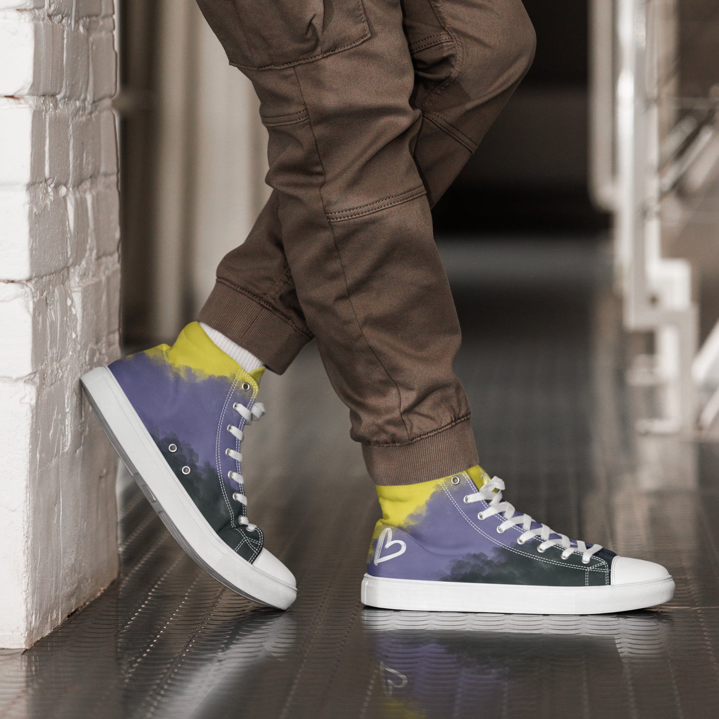 A model wears joggers and a pair of high-top canvas shoes with cloudy color blocks of the yellow, purple, and black non-binary flag colors with white laces and accents, a white heart on the heel, and white Aras Sivad Studio logo on the tongue.