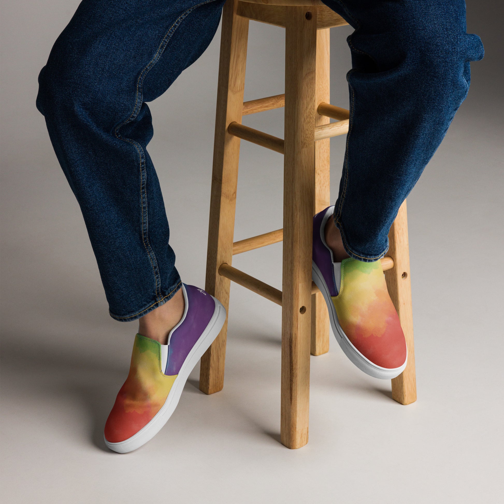 A model wears the cloudy rainbow pride slip on shoes and sits on a stool.