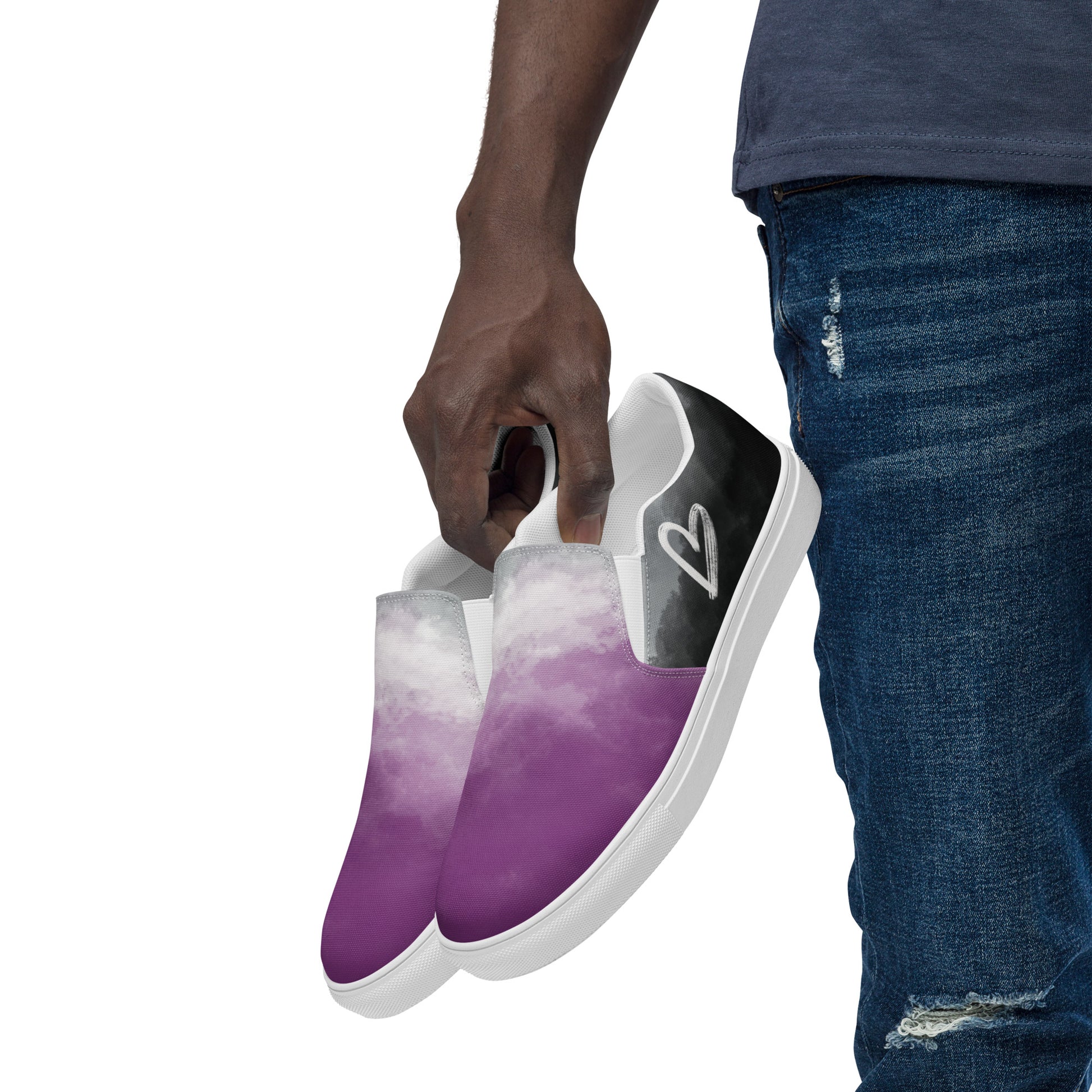 A model holds a pair of slip-on shoes with clouds in the asexual flag colors, a hand drawn white heart on the side, and the Aras Sivad Studio logo on the back.