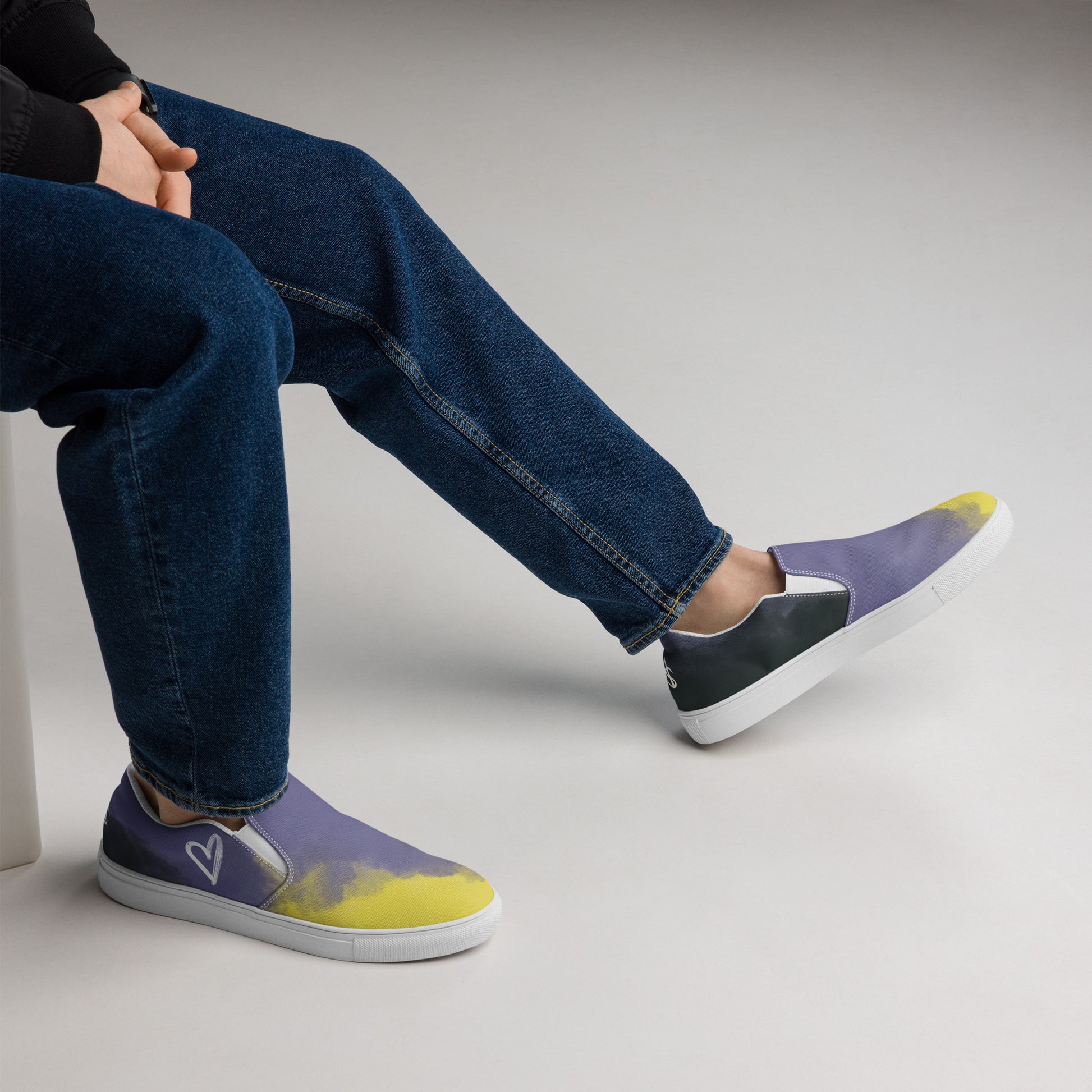 A model wears a pair of slip-on shoes with the non-binary colors in wisps of clouds with a white hand drawn heart on the outside under the ankle and the Aras Sivad Studio logo in white on the back.