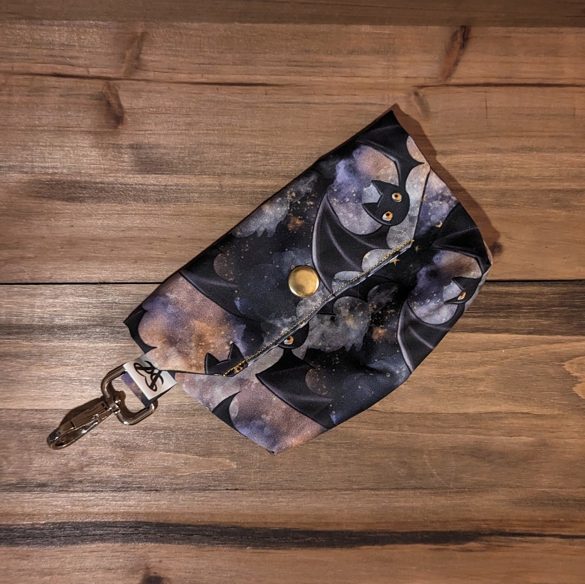 A tarot card case with cute bats flying in purple, amber, and gold clouds outside, a gold snap, and a keychain clip.