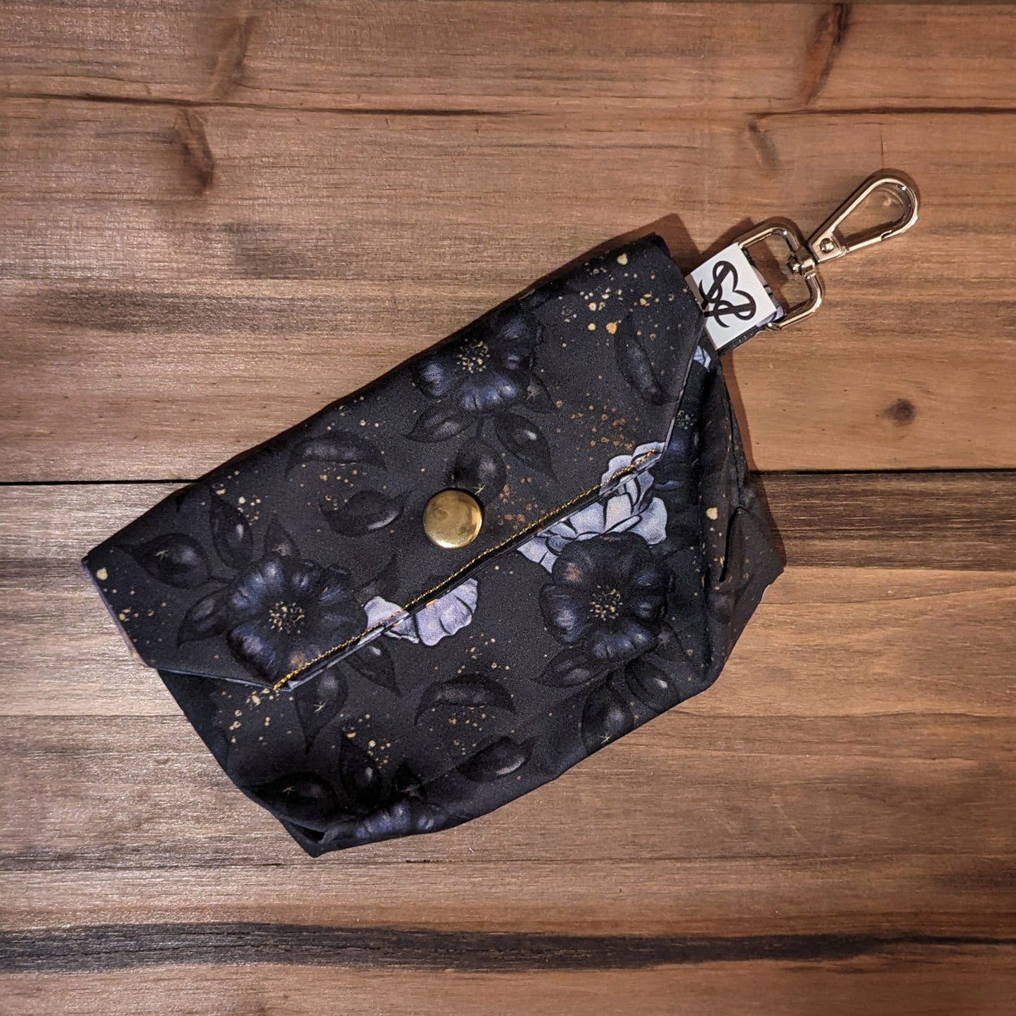 A tarot card case with black and lavender roses, black leaves, and gold speckles outside, a gold snap, and a keychain clip.