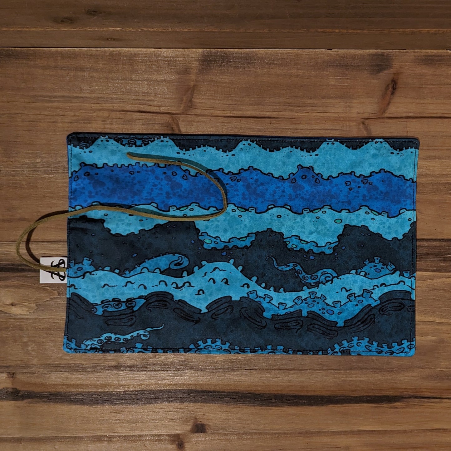 The outside of the Ominous Sea mini utility roll, showing a tentacle and sea foam striped pattern in shades of blue and teal with a green leather cord.