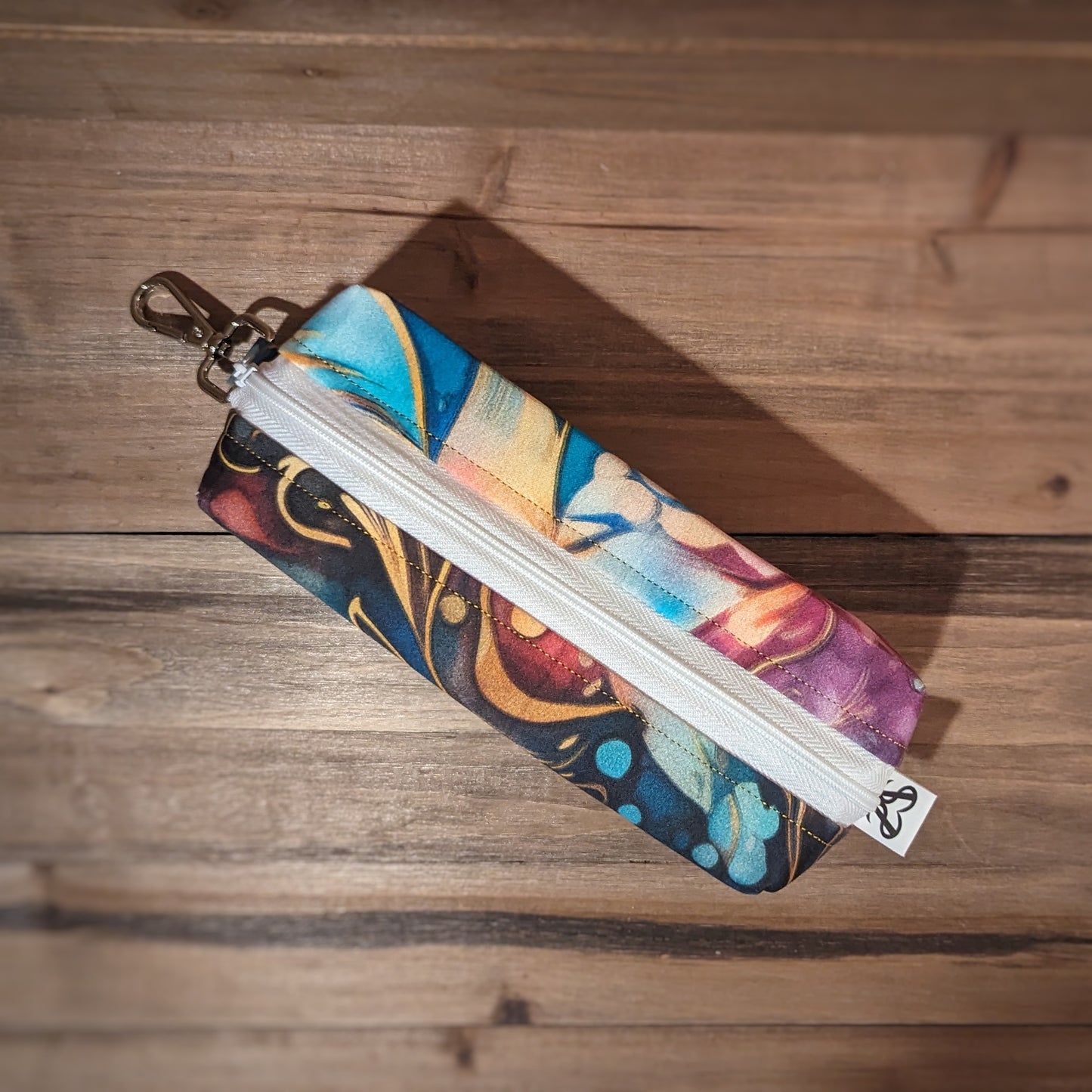 A wide square case with swirling rainbow colors outside and a white zipper up the middle and a keychain clip at the top.