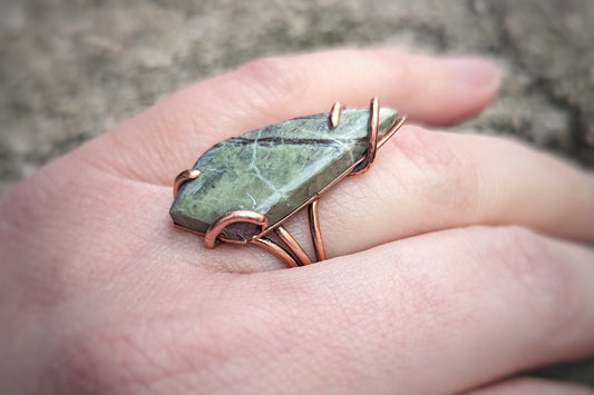 Aras models a copper ring with a skinny kite shaped green Atlantisite stone held on by swirling asymmetrical prongs.