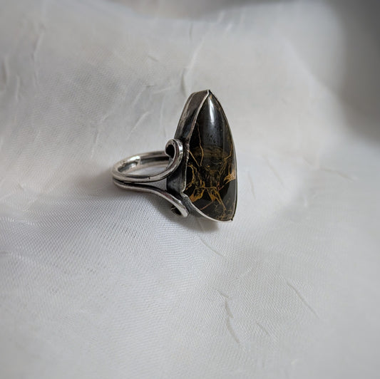 A black triangular stone with orange swirls is set in silver with the two wire ring shank extending up the side to form a swirling frame on one corner.