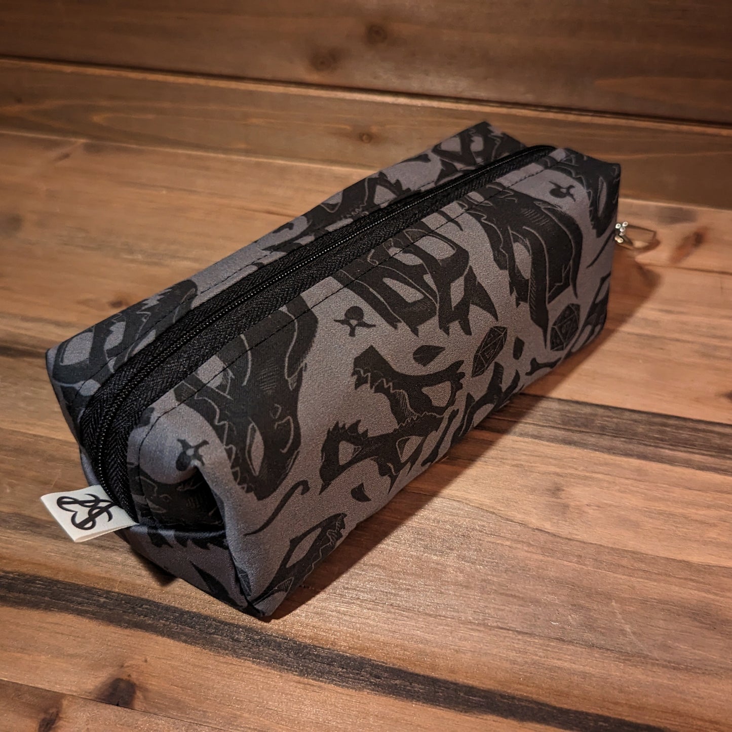 A 7 inch long bag has 2.5 inch square ends with black and grey dragon skull and D20 print fabric on the outside, and a black zipper running long ways from the keychain clip at the top to the Aras Sivad Studio logo at the bottom.
