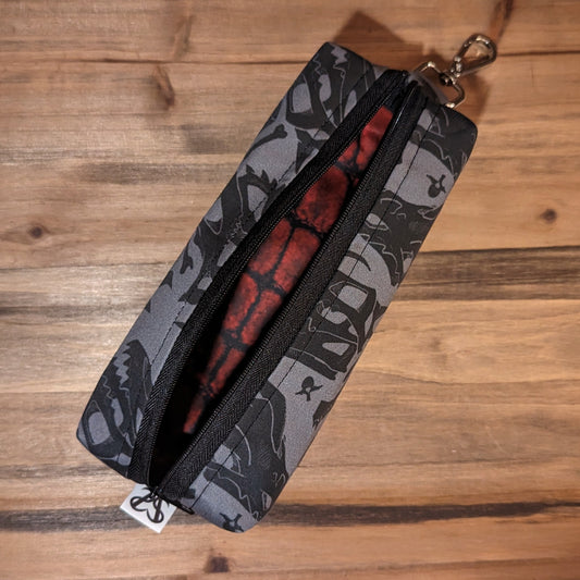 A 7 inch long bag has 2.5 inch square ends with black and grey dragon skull and D20 print fabric on the outside, and a black zipper running long ways, open to show the deep red scale pattern inside.