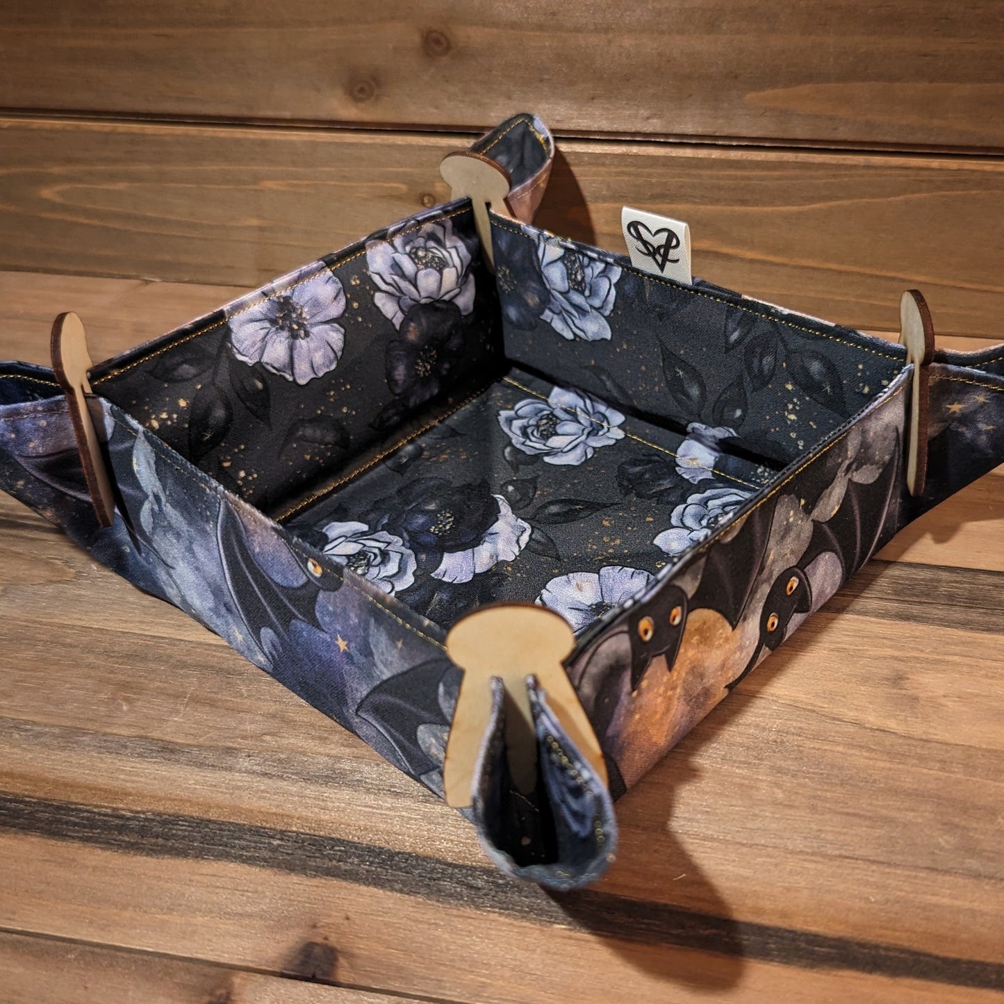 A fabric tray has cute bats flying all over the outside and a bed of black and lavender roses in gold speckles inside, gold thread, and wood clips in the corners.