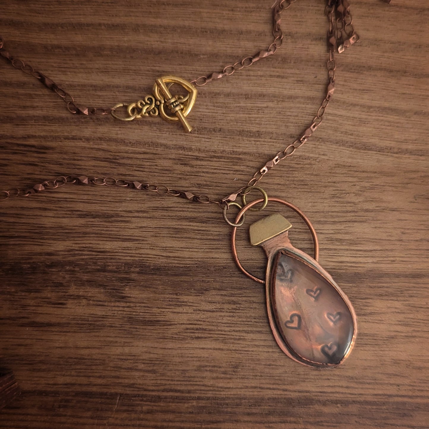 A copper necklace with brass accents is a delicate hanging potion bottle with a rose quartz set over stamped hearts like little bubbles inside and a heart toggle clasp.